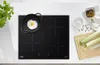 A black induction hob with a frying pan and egg on it, next to leeks and spices on a cutting board. Produktbild-Vorschau