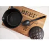 A cast iron skillet, a spoon, and a slotted spoon are arranged on a wooden cutting board with the word "BEEF" and an illustration of a cow. A white plate is partially visible in the foreground. Náhled obrázku produktu