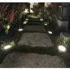 A pathway made of large stone slabs is illuminated by small solar lights embedded in the grass on either side. Lush green grass and flowering plants line the path, creating a serene garden atmosphere. Anteprima dell’immagine prodotto