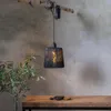 A black rattan-look pendant lamp hangs from a branch, casting a warm glow. A wooden table with stones sits in the foreground, and a plant is to the left. Produktbild-Vorschau