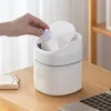 A hand is dropping a crumpled piece of paper into a small white trash can on a wooden desk. Produktbild-Vorschau