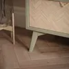 Close-up of the bottom corner of a cabinet with a herringbone wood pattern and green legs, resting on a wooden floor. Produktbild-Vorschau