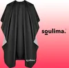 A black hairdressing cape with gray accents is displayed against a pink and white gradient background. The brand logo 'soulima.' is visible on the right side of the image and also on the bottom left of the cape. Anteprima dell’immagine prodotto
