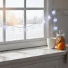 A cozy winter scene with snowflake lights strung across a window, a steaming mug of coffee, cookies, and an orange on a countertop. Náhled obrázku produktu