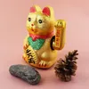 A golden Maneki-neko cat figurine sits on a pink background, with a rock and a pinecone placed in front of it. Produktbild-Vorschau