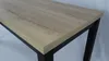 A close-up view of a table with a light brown wood-grain tabletop and black legs. Náhled obrázku produktu