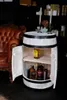 A wooden barrel-shaped bar cabinet is open, displaying bottles of alcohol inside. A leather armchair is visible to the left. Produktbild-Vorschau