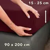 Hands adjusting a burgundy fitted sheet on a mattress. Produktbild-Vorschau