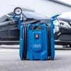 A blue Scheppach air compressor with attached hoses and tools is placed on a paved surface in front of a dark car. Produktbild-Vorschau