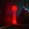 A tall, cone-shaped lamp with a pleated shade, illuminated with red light, stands on a paved area next to a dark, slatted wall and some greenery. Produktbild-Vorschau