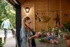 A woman is arranging flowers on a table in a wooden shed, while a man and a dog walk by outside. Anteprima dell’immagine prodotto