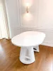 A white oval dining table with a slatted base is shown in a room with white paneled walls and a light fixture. Voir les images du produit