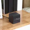 A black digital alarm clock with a white LED display showing the time sits on a wooden surface, with books in the background. Produktbild-Vorschau