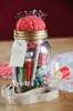 A sewing kit organized in a glass jar, with a pincushion on top, placed on a wooden surface with fabric and spools of thread in the background. Voir les images du produit