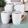 Four white ceramic mugs with a textured honeycomb pattern and a dark rim are arranged on a table with a plant in the background. Podgląd zdjęcia produktu