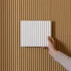 A hand holding a white decorative panel against a fluted brown wall. Produktbild-Vorschau