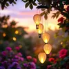 A solar-powered wind chime with five balloon-shaped lights hangs from a tree branch, illuminated by the warm glow of sunset and surrounded by blooming flowers. Produktbild-Vorschau