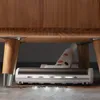 A cordless vacuum cleaner head with LED lights is cleaning under a wooden table. Náhľad obrázku produktu