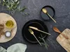 A table setting with black plates, gold cutlery, bread, olive oil, salt, and a wooden spoon on a dark stone surface, with greenery. Náhľad obrázku produktu
