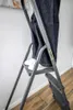 A person is standing on a gray metal step stool, indoors, next to a white wall and a wooden floor. Voir les images du produit