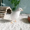 A white quail stands on a sandy substrate in an enclosure with a wooden tunnel and a plant. Produktbild-Vorschau