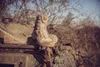 A pair of beige tactical boots are placed on top of a rusty, weathered metal surface, possibly part of a military vehicle, with a blurred background of trees and a clear sky. Produktbild-Vorschau
