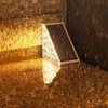 Weiße dreieckige Solar-Bodenleuchte mit Solarmodul, beleuchtet und reflektierend auf nasser, strukturierter brauner Oberfläche im Regen Náhľad obrázku produktu