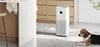 A white air purifier is placed on a speckled floor next to a beagle dog, with kitchen cabinets and a counter in the background. Produktbild-Vorschau