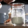 A glass jar with a clamp lid filled with powder sits on a wooden surface next to a blue mug and some spilled flour. Hands are visible in the background, suggesting preparation. Produktbild-Vorschau