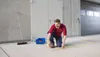 A man is kneeling on the ground, using a yellow measuring tape to mark a rectangular area on the concrete floor. A broom and a blue toolbox are nearby. Náhled obrázku produktu