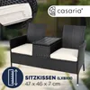 A black rattan garden bench with white cushions and a central table, shown outdoors with a blurred background. Produktbild-Vorschau