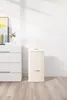 A cream-colored trash can with a silver handle is placed on a light wooden floor next to a white dresser. A painting with yellow and blue abstract art hangs on the wall behind the dresser. Voir les images du produit