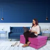 A woman is sitting on a pink floor cushion with a laptop, in a room with blue walls and a blue couch. Podgląd zdjęcia produktu