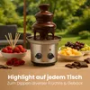 A chocolate fountain is set up outdoors on a table with various fruits and chocolates for dipping. Podgląd zdjęcia produktu
