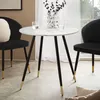 A round marble-look dining table with black and gold legs, placed between two black chairs in a room. Produktbild-Vorschau