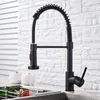 A black kitchen faucet is shown in use, with water running into a sink. The background features a white brick wall and a white slatted blind. Náhľad obrázku produktu