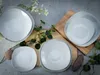 A top-down view of a set of square white plates and bowls arranged on a textured surface with some greenery and decorative elements. Náhľad obrázku produktu