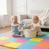 Two babies playing with stuffed animals on a colorful puzzle mat in a living room. Produktbild-Vorschau