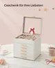 A white jewelry box with multiple drawers and a glass lid is open, displaying various jewelry items. A small heart-shaped decoration with a red ribbon is attached to the inside of the lid. The box sits on a marble-like surface, with decorative elements lik Produktbild-Vorschau