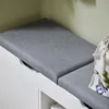 A close-up view of a white storage bench with a gray upholstered seat, showing a portion of a bag with green beads on the right. Produktbild-Vorschau