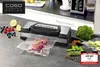 A vacuum sealer is shown on a kitchen counter, sealing a piece of meat. Various fresh ingredients like berries, lemon, broccoli, and zucchini are arranged around it. Anteprima dell’immagine prodotto