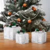 Three illuminated Christmas gift boxes are arranged on a wooden surface in front of a blurred Christmas tree. Anteprima dell’immagine prodotto