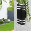 A modern black outdoor wall light mounted on a white brick wall, with a garden in the background. Náhľad obrázku produktu