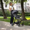 A woman is holding a baby while standing next to a stroller on a paved path in a park-like setting with trees and a building in the background. Náhľad obrázku produktu