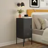A black metal nightstand with a locked door and ventilation slots sits next to a bed with a yellow headboard and patterned pillows. A small plant in a pink vase and a camera on top of books are on the nightstand. A wall lamp is mounted above. Anteprima dell’immagine prodotto