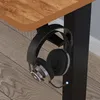A close-up view of a pair of black headphones hanging from a black headphone stand attached to the underside of a wooden desk. Voir les images du produit