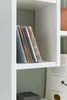 A close-up view of a white shelf with several CDs and a book on it. The background is a light green wall. Produktbild-Vorschau