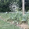 A kinetic wind spinner is placed in a garden bed with lush green plants. Anteprima dell’immagine prodotto