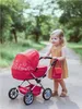 A young girl is pushing a red doll stroller on a path in a park. Náhľad obrázku produktu