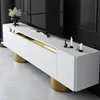 A white TV stand with gold accents and cylindrical gold legs, placed on a light-colored floor. A large black television is visible in the background. Náhľad obrázku produktu